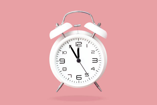 istock White bell alarm clock hovering over pastel pink background. Clock face 5 to 12 1300505789