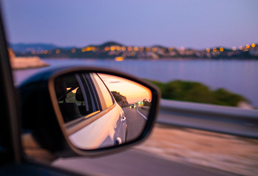 Traveling by car along beautiful coast at night. Reflection of sunset at side mirror