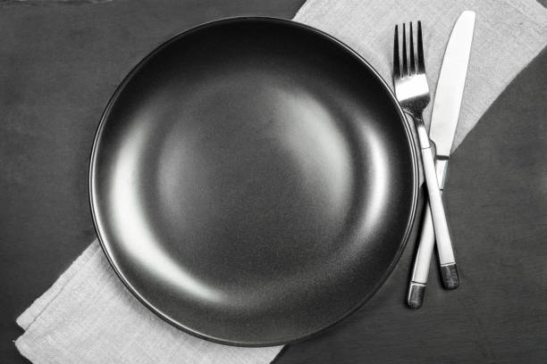 istock Empty blank black plate and cutlery on grey diagonal cooking towel on black shale serving board. 1300507997