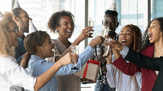 Overjoyed diverse multiracial businesspeople have fun celebrate employee birthday clink glasses in office. Happy multiethnic colleagues laugh have celebration cheers at party at workplace together.