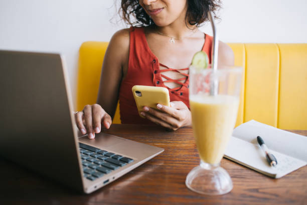 cropped image of skilled woman using cellphone gadget and laptop computer for sharing media files during bistro visiting, female freelancer with mobile phone writing message during netbook chatting - blueberry smoothie milk shake drink imagens e fotografias de stock