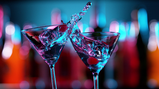 Closeup of splashing martini cocktails in cheers gesture. Bar on background, free space for text