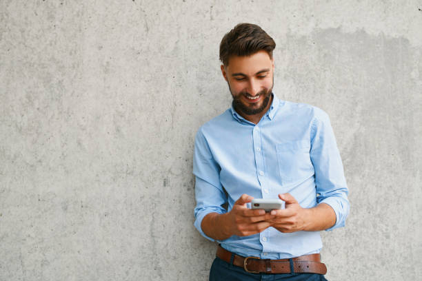 istock Smiling young man leaning against wall and using phone 1300525603