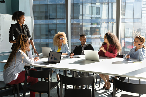 Young African American businesswoman lead meeting with multiethnic colleagues, communicate interact in office. Female ethnic coach or boss present at workplace, brainstorm with diverse businesspeople.