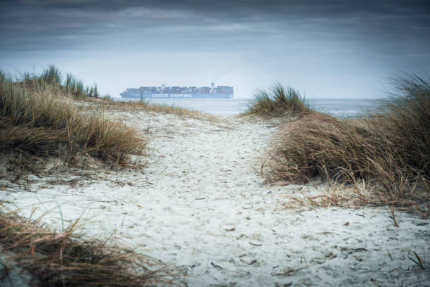 Close-up view of the dark, rough sea with a breaking white wave and a large cargo ship on the distant horizon. The dramatic contrast between the turquoise water and the heavy, black storm clouds creates a sense of imminent weather change. Photographed near Skagen, Denmark.