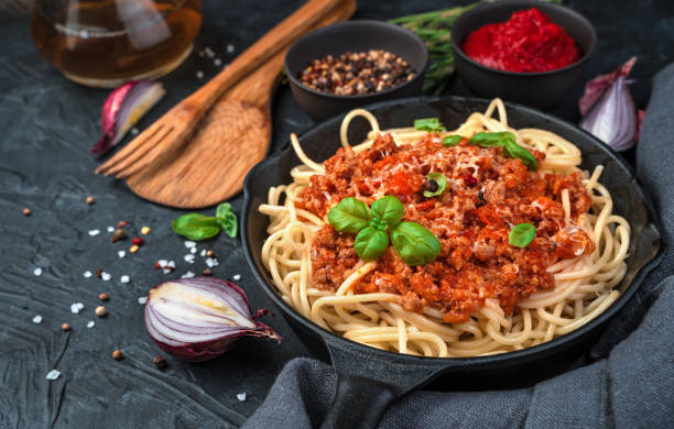 spaghetti and sauce in a frying pan are sprinkled with cheese and fresh basil on a background of spices and onions on a black concrete background - spaghetti imagens e fotografias de stock