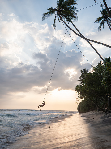 Rope swing holding on tropical palm tree , female enjoying sunset swing above beach