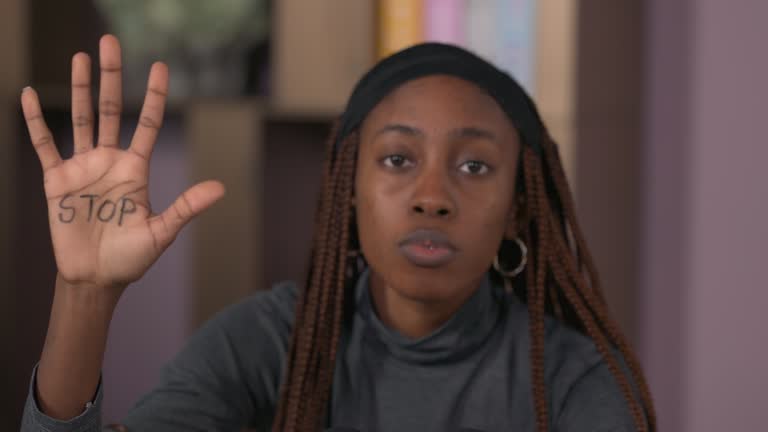 Close up of African young woman sitting on floor in room, holding knees with arms and raising palm with stop inscription. Women against domestic violence. Tilting camera.