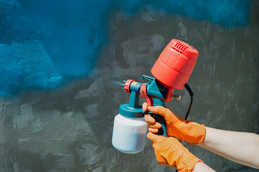 Painting a roughly plastered wall with a hand-held spray gun. Spray gun without hose with overhead compressor.A worker's hand in a yellow glove holds a hand spray gun with an overhead compressor. Painting a roughly plastered wall blue. Household renovation. Copy space. Selective focus
