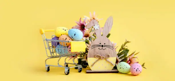 Photo of Easter composition with colorful eggs in shopping cart, wooden bunny and spring flowers on yellow background. Banner.