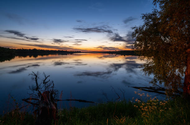 istock Summer evening twilight view on picturesque plain lake with forest and blossoming meadows on shores and sunset clouds reflections in water surface. 1300559551
