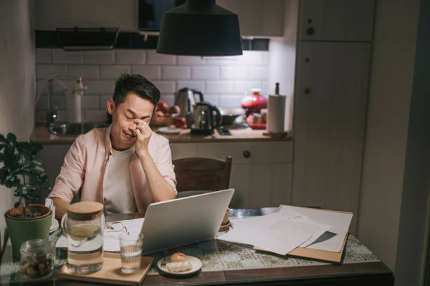 night overwork Asian chinese male working late at home dining room emotional stress with his financial bill alone at night Asian chinese male working late at home dining room emotional stress with his financial bill alone at night debt stock pictures, royalty-free photos & images