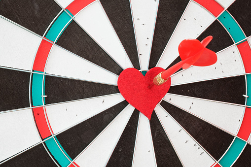 Dart arrow on red heart shape in on the bull's eye of dartboard. Pixel shift effect is applied for more details. No people are seen in frame. Shot with a full frame mirrorless camera.