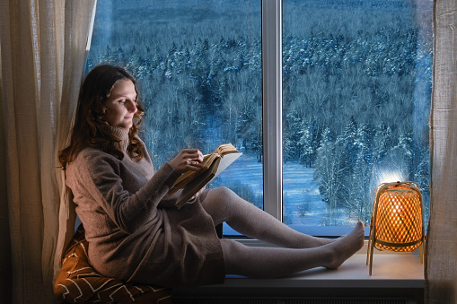 Young woman reading a book by the window in the light of the evening lamp. Woman sitting on the windowsill by the window with snow-covered trees in the winter forest