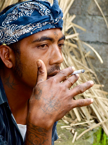 Closeup portrait of a young Balinese man sitting in his family compound in a small country village in Bali, smoking a cigarette. He wears a traditional male batik head bandana, patterned blue and white.