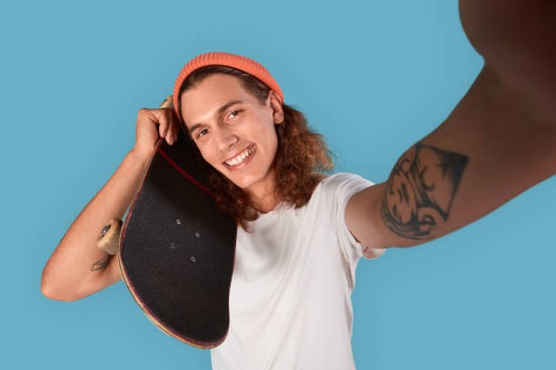 istock Freestyle. Curly-haired guy wearing hat standing isolated on blue with skateboard taking selfie smiling cheerful close-up 1300582926
