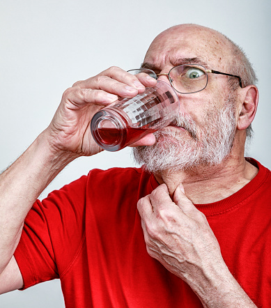 A humorous, grimacing, very distressed senior adult man apparently hates taking his medicine pills. He's trying to swallow a tiny medication tablet he's just put in his mouth by attempting to wash, will, and force it down his throat with the combination of a frantic gulp of cranberry fruit juice from the drinking glass he is holding, and a panicked grab of his neck to try to stretch his throat.