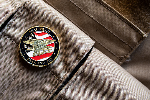 A coin issued to certain people by the US Navy resting atop a magazine pouch of a plate carrier background.
