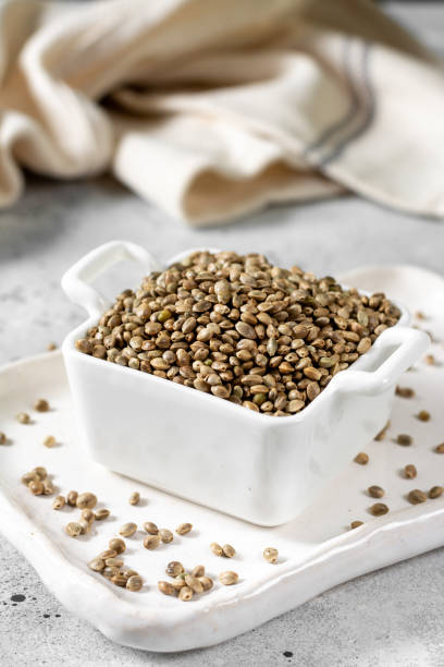 hemp seeds. peeled and whole dried hemp seeds in a white ceramic bowl on a light gray kitchen table. hemp seeds for cooking - hemp seed heap white imagens e fotografias de stock