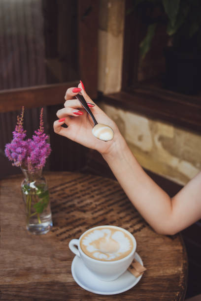 la mano de una mujer sostiene una cuchara con espuma de leche de la taza de café en una mesa de madera - women spoon tasting elegance fotografías e imágenes de stock