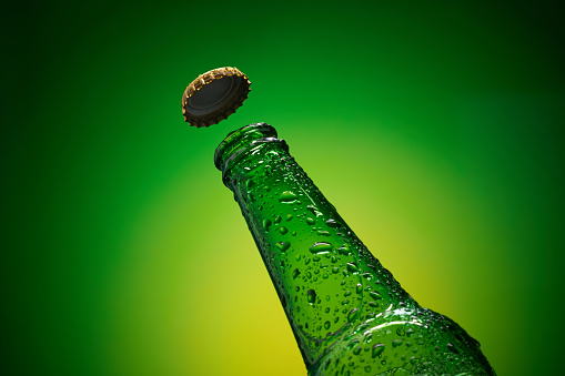 Green cold bottleneck with drops and flying golden cap against yellow green background. High resolution, high post-production quality.