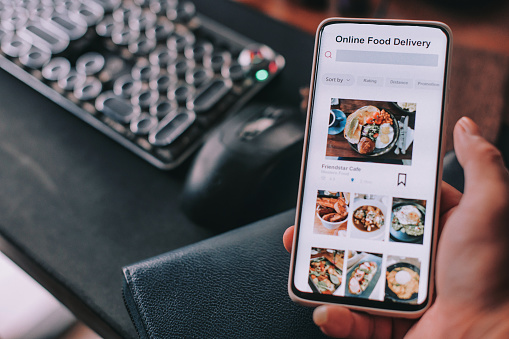 Online food delivery mobile app shown on smart phone screen hold by asian man hands in front of desktop pc