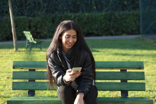 istock Young woman sitting on a bench in a public park using smartphone 1300662228