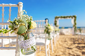 Luxurious wedding ceremony on the ocean, beach. White chairs decorated with a beautiful bouquet of flowers in a jar hanging on them.