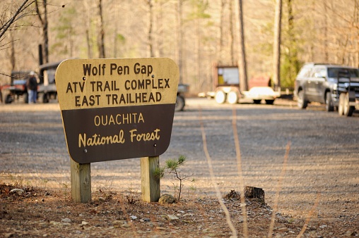 Sign in Ouachita National Forest for Wolf Pen Gap ATV Trail Complex East Trailhead with cars  and ATV trailers parked in background