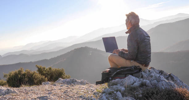 mature man uses computer on mountain top at dawn - escape from it all fotos imagens e fotografias de stock