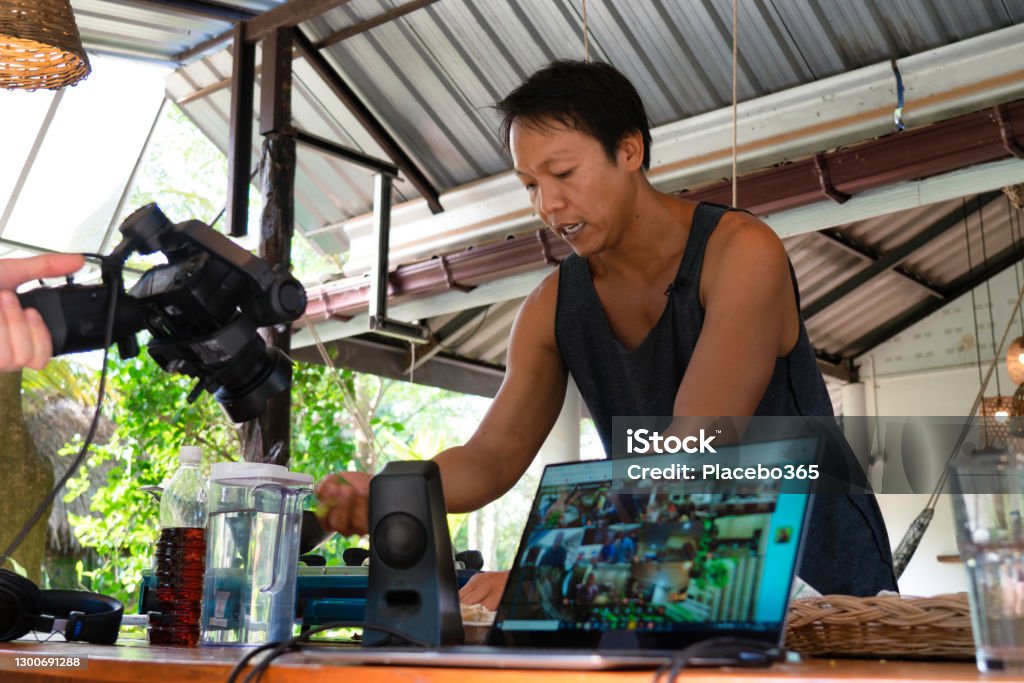 Asian Youtuber live streaming from home A Thai Youtuber is live streaming a tutorial from home.  The camera operator films him and broadcasts the live event. Live Streaming Stock Photo