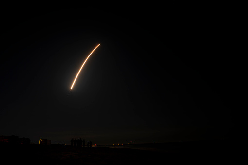 A rocket launches at night from Cape Canaveral, Florida, at 1:19 a.m. on February 4, 2021. A small group of people are in the foreground, standing on the beach to watch the launch. Photo taken in Cocoa Beach using a 30-second exposure.