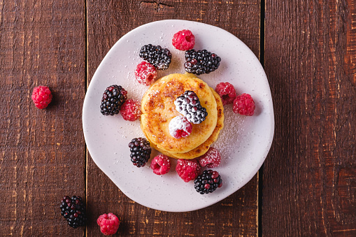 Cottage cheese pancakes and powdered sugar, curd fritters dessert with raspberry and blackberry berries in plate on dark brown wooden background, top view
