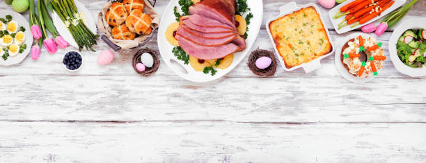 istock Traditional Easter ham dinner. Overhead view top border on a white wood banner background with copy space. 1300699582