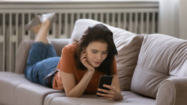 istock Concentrated millennial female lying on couch on belly using smartphone 1300701395