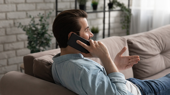 Talking on phone. Calm young man resting on couch at home holding phone to ear making answering audio call. Relaxed male having nice talk with wife girlfriend or ordering food at delivery service