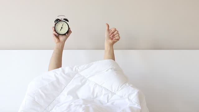 Man showing arm raised up black alarm clock behind duvet in the bed room, Young boy with two hands sticking out from the blanket. wake up with fun in morning concept.