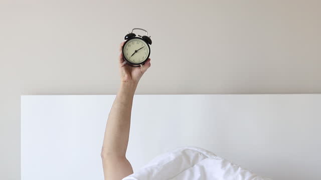 Man showing arm raised up black alarm clock behind duvet in the bed room, Young boy with hands sticking out from the blanket. wake up with fun in morning concept.