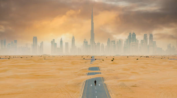 vista dall'alto, splendida vista aerea di una persona non identificata che cammina su una strada deserta coperta da dune di sabbia con lo skyline di dubai sullo sfondo. dubai, emirati arabi uniti. - fog desert arabia sunset foto e immagini stock