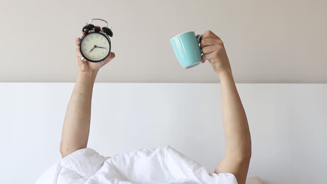 Man showing arm raised up holding coffee cup and black alarm clock behind duvet in the bed room, Young boy with two hands sticking out from the blanket. wake up with fun in morning concept.