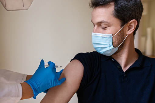 Mid-adult man wearing face mask getting the covid-19 vaccine by a doctor. Doctor wearing protective glove injecting the vaccine into patient's arm in the clinic.