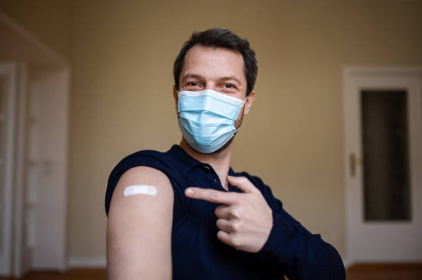Happy to be in first few to get vaccinated Man wearing protective face mask pointing at his arm with a bandage after receiving the covid-19 vaccine. adhesive bandage photos stock pictures, royalty-free photos & images