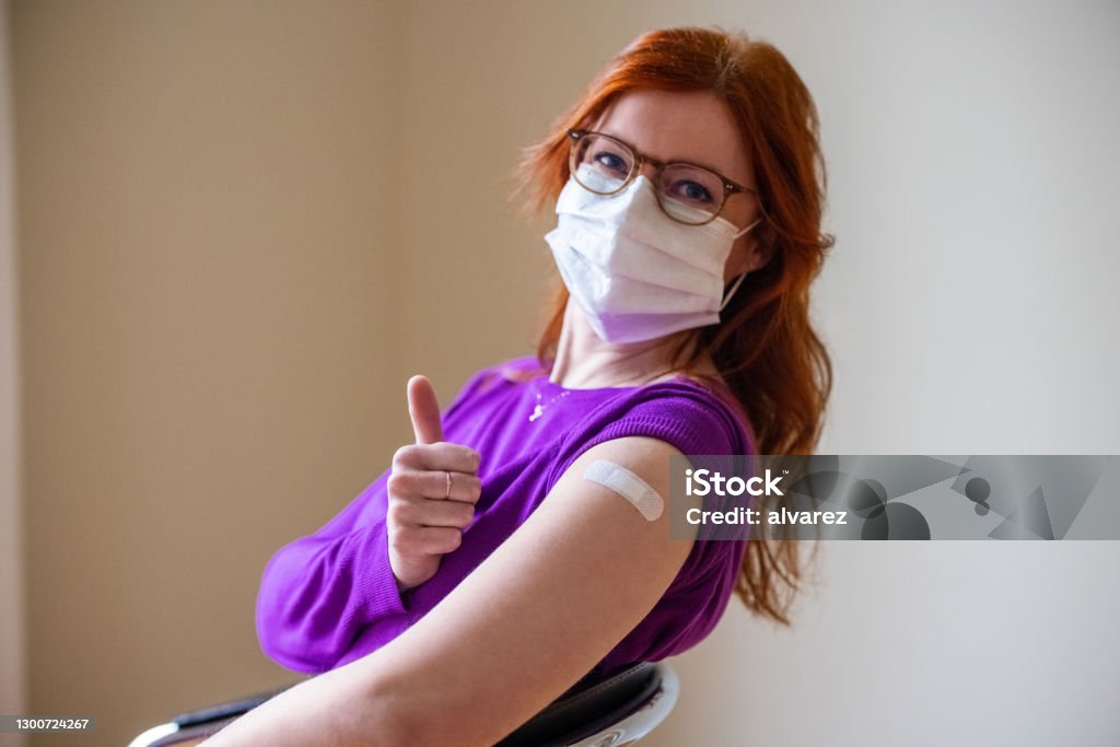 Woman showing thumbs up after getting covid-19 vaccine Woman wearing face mask looking at camera showing thumbs up after getting the covid-19 vaccine. Female gesturing thumbs-up sign after getting coronavirus vaccine at vaccination center. Vaccination Stock Photo