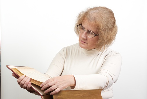 Mature age woman with glasses sits while reading old red paperback book (Bible)