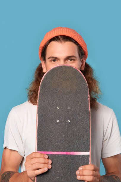 istock Freestyle. Curly-haired guy wearing hat standing isolated on blue covering face with skateboard looking camera curious 1300739581