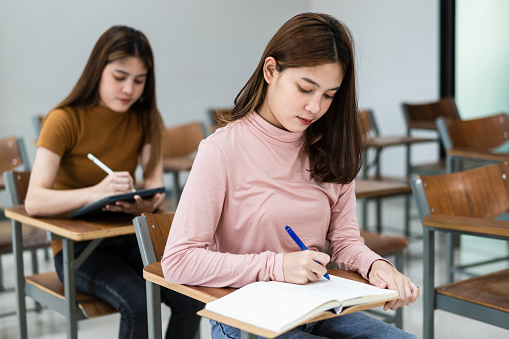 Young woman college students sitting on lecture chair in classroom studying, writing on examination paper answer sheet in doing the final examination test. College students in the classroom present campus life concept.