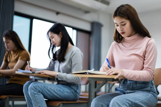 istock Young woman college students sitting on lecture chair in classroom studying, writing on examination paper answer sheet in doing the final examination test. College students in the classroom present campus life concept. 1300771469