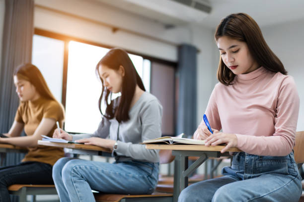 istock Young woman college students sitting on lecture chair in classroom studying, writing on examination paper answer sheet in doing the final examination test. College students in the classroom present campus life concept. 1300771519
