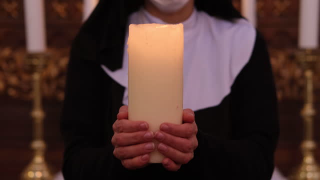 Nun holding a big white candle in Roman Catholic church, wearing protective face mask.