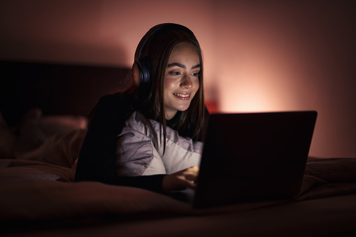 Young teenage woman relaxing on her bed in bedroom at night, watching streaming videos - wearing overear headphones on her Laptop Computer. Natural Ambient Night Bedroom Light. Millenial Generation Modern Technology Daily Lifestyle.